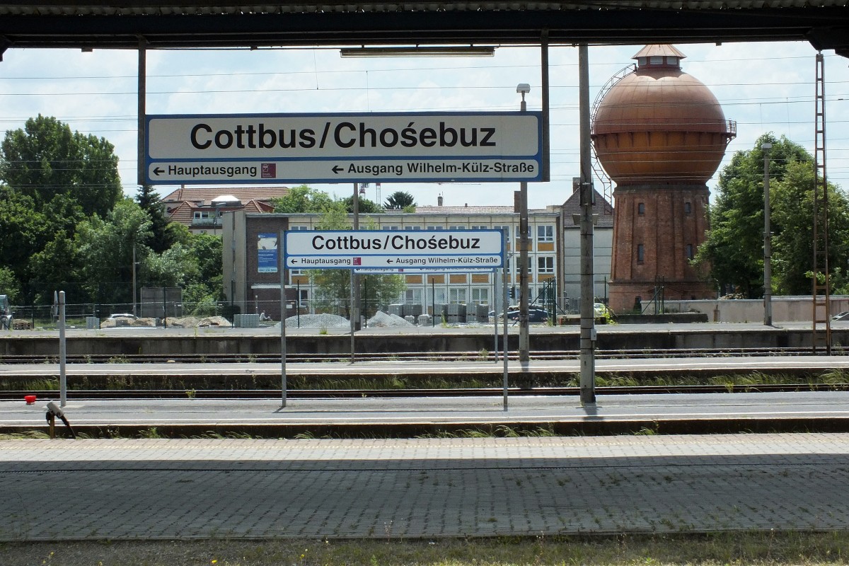 Cottbus. Wir befinden uns im Land der Sorben, neben Friesen, Dänen und Roma eine von vier offiziell anerkannten Nationalen Minderheiten in der Bundesrepublik Deutschland. In weiten Teilen Brandenburgs und Sachsen sind Straßenschilder oder, wie hier zu sehen, Bahnhofsschilder deshalb zweisprachig ausgeführt: Einmal hochdeutsch, einmal sorbisch.
Muttersprachlich gesprochen wird die Sprache heute kaum noch, historisch ist sie aber immer noch bedeutsam genug, um sie allgegenwärtig zu halten. Nicht zuletzt weil Sprache immer eine wichtige Identitätsstiftung war und ist. Bahnhofsszene in Cottbus HBF, wo noch immer die weißen Stationsschilder hängen, die ich sonst in keinem Bahnhof auf meiner Tour sichten konnte, 30.5.14